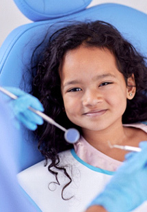 Girl with long hair in dental chair smiling as dentist approaches with instruments for exam